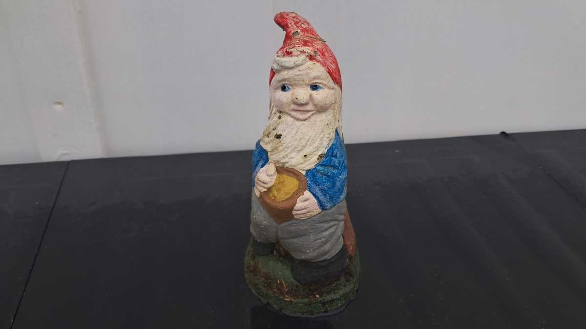The little dude gnome is wearing a blue shirt and grey pants. He is sitting on a brown treestump and holding a pot of gold.